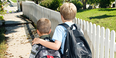 Two young boys wearing backpacks, walking down a sidewalk. The taller boy has his arm protectively over the smaller boy's shoulders.