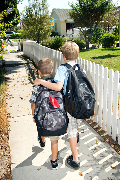 Two young boys wearing backpacks, walking down a sidewalk. The taller boy has his arm protectively over the smaller boy's shoulders.